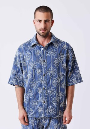 Homme aux cheveux courts portant une chemise boutonnée bleue et un pantalon assorti avec des motifs floraux blancs en ligne, debout devant un fond uni.