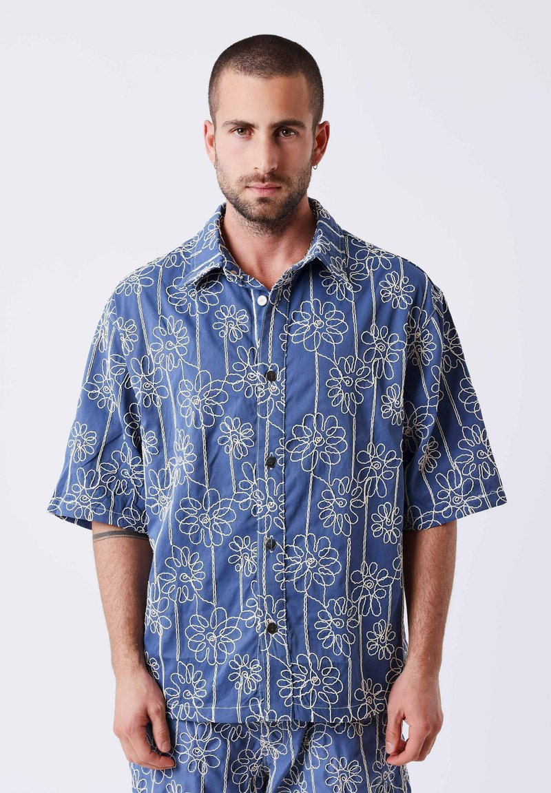 Homme aux cheveux courts portant une chemise boutonnée bleue et un pantalon assorti avec des motifs floraux blancs en ligne, debout devant un fond uni.