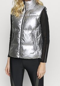 Silver pufferväst med hög krage, dragkedjeöppning och två sidofickor. Har en glänsande yta och svart Champion-logotyp på bröstet.