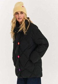 Czarna pikowana kurtka typu puffer z wysokim kołnierzem i kolorowymi guzikami w odcieniach różu, pomarańczy i fioletu. Nosi ją z żółtą dzianinową czapką.