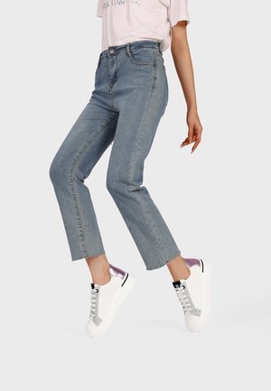 Donna che indossa jeans corti azzurri e sneakers bianche con tacchi metallici viola, a metà passo su uno sfondo neutro.