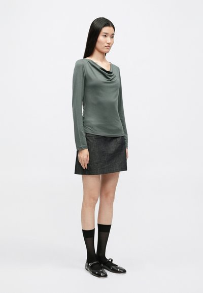 Camisola de manga longa com gola alta em tecido verde suave, combinada com uma mini saia de ganga escura. Meias altas pretas e sapatos planos com detalhes.