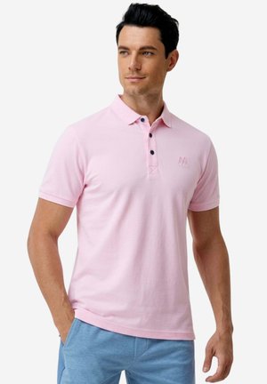 Mann mit dunklen Haaren, der ein hellrosa Polohemd mit drei Knöpfen und hellblaue Hose trägt, steht mit einer Hand in der Tasche.