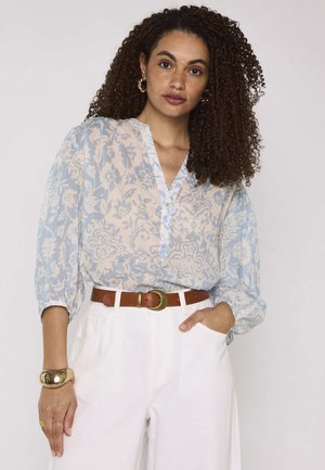 Femme aux cheveux bouclés porte un chemisier bleu clair à fleurs, un pantalon blanc, une ceinture marron, un bracelet doré et des créoles, debout avec une main dans la poche.