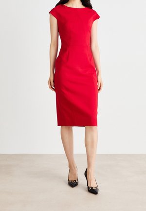 Kobieta ubrana w dopasowaną, czerwoną sukienkę sięgającą do kolan z krótkimi rękawami i czarne szpilki ze złotymi akcentami, stojąca na jasnej podłodze.