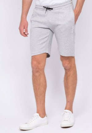 Homme portant un short gris clair à cordon et des baskets blanches, debout sur un fond blanc.