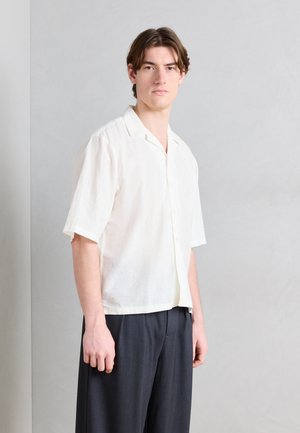 Junger Mann mit braunen Haaren trägt ein kurzärmeliges weißes Hemd mit Knöpfen und dunkle, lockere Hose, steht vor einer schlichten hellgrauen Wand.