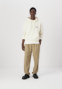Haut à capuche couleur crème avec cordons, logo bleu sur la poitrine ; associé à un pantalon plissé beige et des chaussures noires à semelles épaisses. Coupe simple et décontractée.
