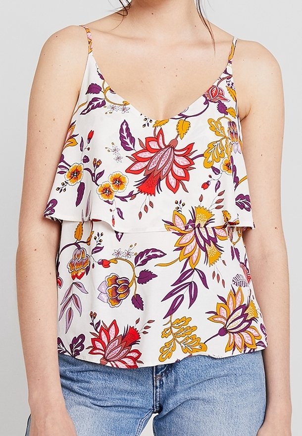 Femme portant un haut sans manches blanc avec un imprimé floral en couches rouge, jaune et violet, associé à un jean bleu.