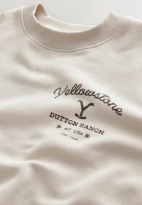 Sweat-shirt beige avec un col rond côtelé. Présente un texte imprimé foncé indiquant "Yellowstone Dutton Ranch MT USA EST. 1886" en arc de cercle.