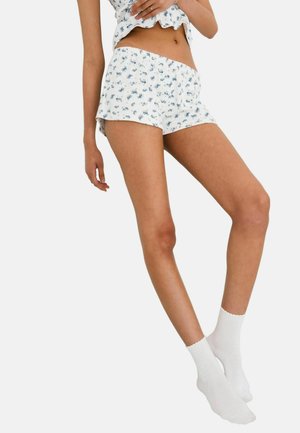 Vrouw draagt witte pyjamashort met bloemmotief en bijpassend shirt, witte enkelsokken, staand tegen een effen achtergrond.