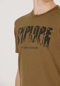 Ruskea puuvillainen t-paita lyhyillä hihoilla, jossa on suuri musta graafinen printti puista ja teksti "TUTKI SUURTA ULKOILMAA."