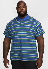 Dryžuotas polo marškinėliai mėlynos, žalios ir rudos spalvų. Turi klasikinį apykaklę ir du mygtukus, o krūtinėje yra balta Nike logotipas. Trumpi rankovės.