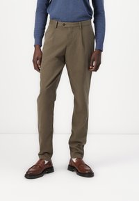 Olivgrüne Hosen mit geradem Schnitt, aus Baumwollstoff. Verfügt über einen einzelnen Knopfverschluss und Vorderfalten. Getragen mit braunen Slippern.