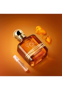 Flacon de parfum Emporio Armani couleur ambre avec bouchon doré et petit flacon vaporisateur à côté de pierres d'ambre sur un fond orange chaud.