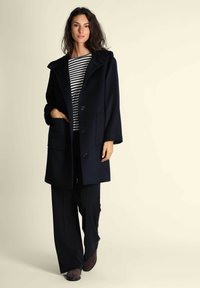 Cappotto in misto lana blu navy con cappuccio oversize, caratterizzato da grandi bottoni e tasche laterali, indossato sopra un top a righe e pantaloni a gamba larga.