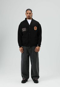 Zwarte cardigan met twee steekzakken aan de voorkant, geborduurd met "23" en een embleem, gecombineerd met loszittende donkergrijze jeans en zwarte schoenen.