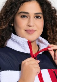 Veste doublée en polaire avec une fermeture éclair rouge, corps marine et accents rayés blancs. Col haut, texture douce et fermeture éclair à l'avant.