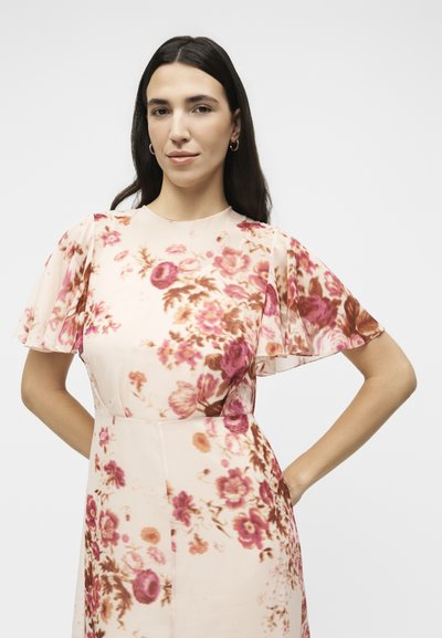 Γυναίκα με μακριά σκουρόχρωμα μαλλιά, φοράει ένα κοντομάνικο, ανοιχτό ροζ φόρεμα με κόκκινα και καφέ λουλουδένια σχέδια, στέκεται με τα χέρια στα ισχία.