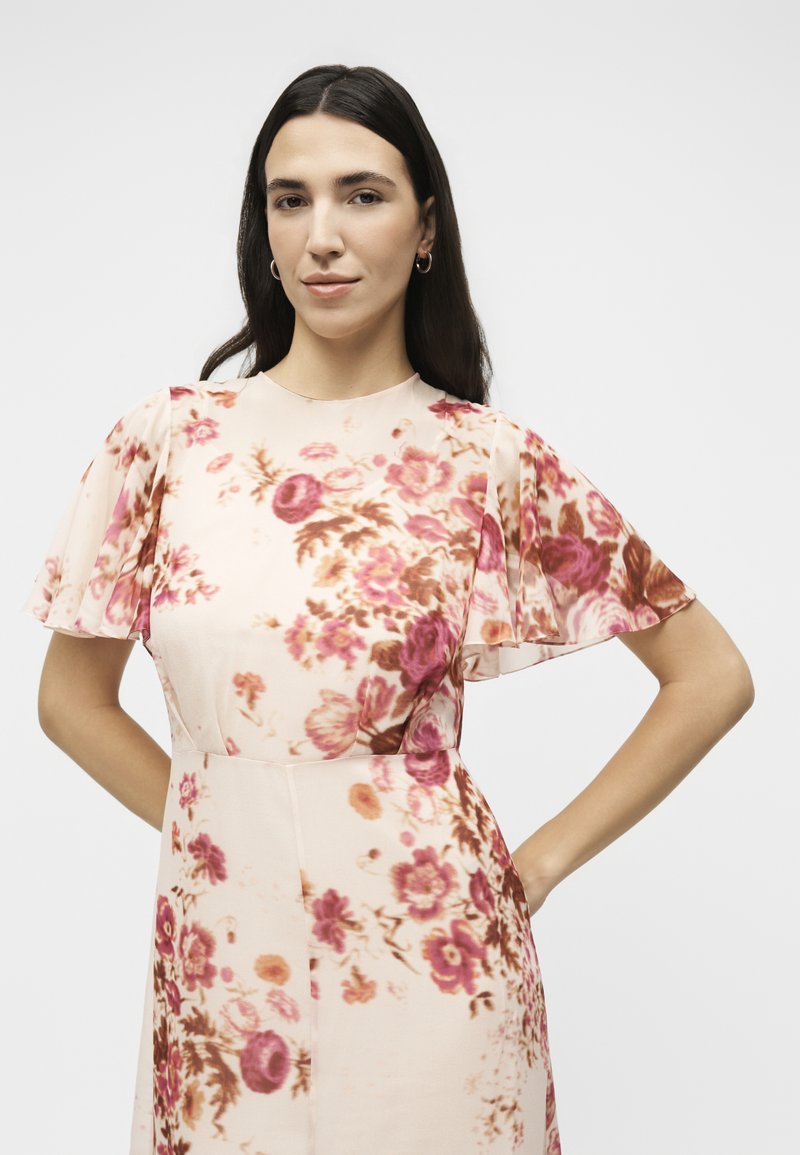 Frau mit langen dunklen Haaren, die ein kurzärmeliges, hellrosa Kleid mit roten und braunen Blumenmustern trägt, steht mit den Händen in der Hüfte.
