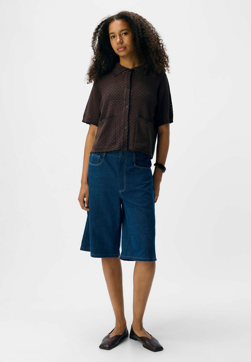 Kobieta z kręconymi włosami, nosząca brązową bluzkę z krótkim rękawem zapinaną na guziki, dżinsowe szorty do kolan, ciemne baleriny i czarną bransoletkę na nadgarstku, stoi w bezruchu.