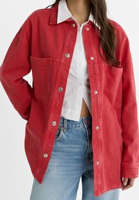 Giacca di denim rossa con chiusura a bottoni, due tasche sul petto e vestibilità rilassata. Abbinata a una camicia bianca con colletto e jeans azzurri chiari.