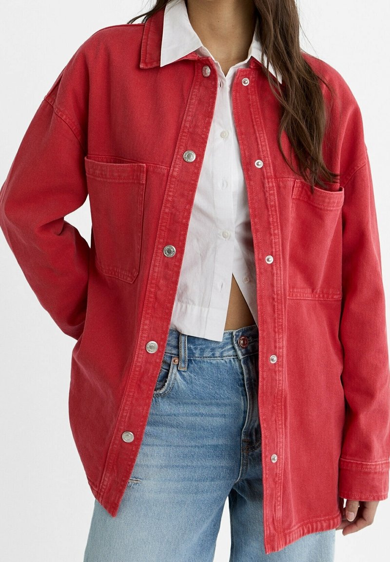 Giacca di denim rossa con chiusura a bottoni, due tasche sul petto e vestibilità rilassata. Abbinata a una camicia bianca con colletto e jeans azzurri chiari.