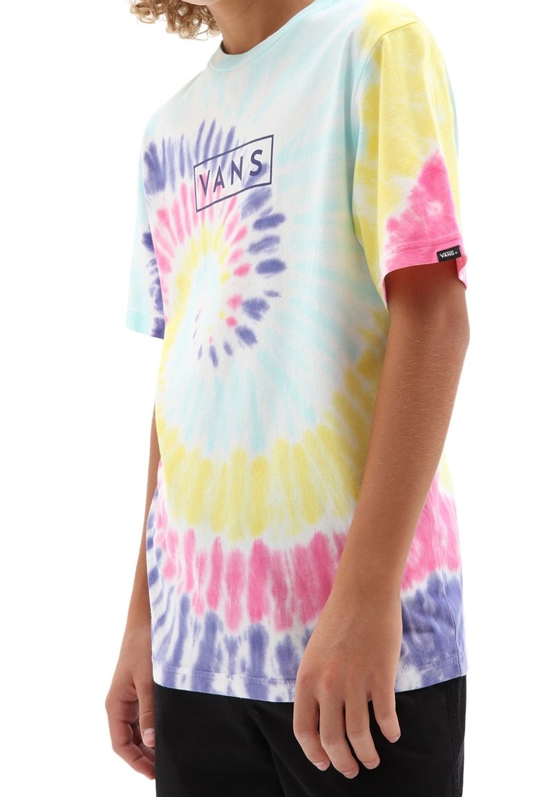 Personne portant un t-shirt tie-dye Vans avec des motifs en spirale jaune, rose, violet et bleu et un pantalon noir, visible du cou jusqu'aux hanches.