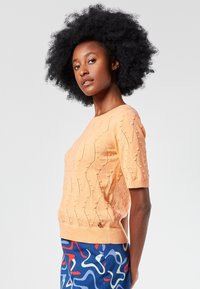 Lyhythiha, persikanvärinen neulepaita, jossa on teksturoitu aaltoileva kuvio ja pompuloita. Ribbauskaulus ja helma, jossa on pieni logoaksentti.