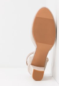 Sandale à talons hauts en daim beige avec une semelle texturée, une bride de cheville ajustable et une large zone pour les orteils, dotée d'un talon bloc stable.