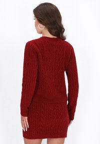 Punainen neulepaita, jossa on kaapelitekstiä, pitkät hihat ja kapea helma. Pehmeä rakenne, jossa on hieno kiilto ja ribatut hihansuut.