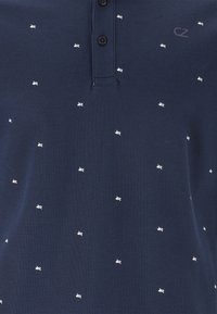 Marineblå pikétröja i strukturerad bomull, med en ståkrage för tre svarta knappar och små vita grafiska mönster över hela.