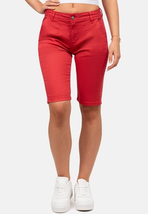 Mujer con pantalones cortos rojos hasta la rodilla, zapatillas blancas y un top blanco corto con una mano en la cadera sobre fondo blanco.