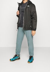 Persona con una chaqueta negra con cremallera, camiseta oscura, pantalones de chándal azul claro y zapatillas deportivas azules y negras, de pie sobre un fondo blanco.