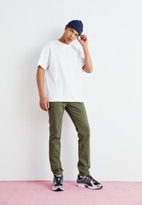 White short-sleeve t-shirt, olive green pants, and black sneakers with red accents. Model wears a navy knit beanie and poses casually.