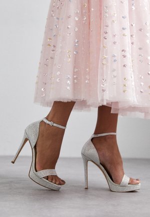 Srebrne sandały na wysokim obcasie z ozdobami z kryształków, z paskiem wokół kostki i odkrytym palcem, zestawione z pastelową spódnicą z cekinami.