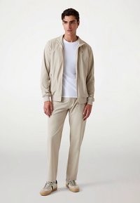 Homokszínű, cipzáras felső és hozzá illő, sima anyagból készült nadrág. Alatta fehér póló. Hétköznapi sportcipők kék és homokszínű részletekkel.