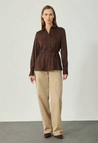 Brown long-sleeve shirt with a button-down front, belted at the waist, paired with beige wide-leg pants and dark shoes. Smooth fabric texture.