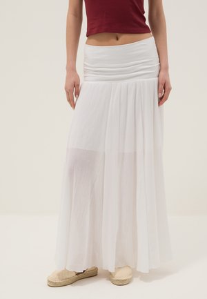 Femme portant une longue jupe blanche fluide avec une taille ajustée, associée à des chaussures slip-on beiges tissées et un haut court bordeaux.