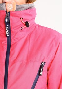Veste imperméable rose avec un col haut, zips navy, cordons élastiques et une douce doublure grise. Comprend une poche latérale avec fermeture éclair.