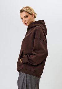 Barna kapucnis pulóver, laza szabással, nagy kenguruzsebbel és bordás mandzsettával. Simább szövet textúra, szürke csíkos nadrággal párosítva.