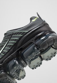 Czarny sportowy but z zielonymi akcentami, teksturowaną cholewką i przezroczystą podeszwą z poduszką powietrzną. Posiada falisty wzór i wzmocniony obcas.