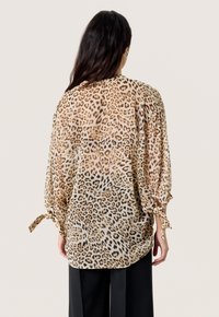 Leopardprint bluse i let, gennemsigtigt stof. Har løs pasform, lange ærmer med bånd og en buet kant. Sorte bukser under.