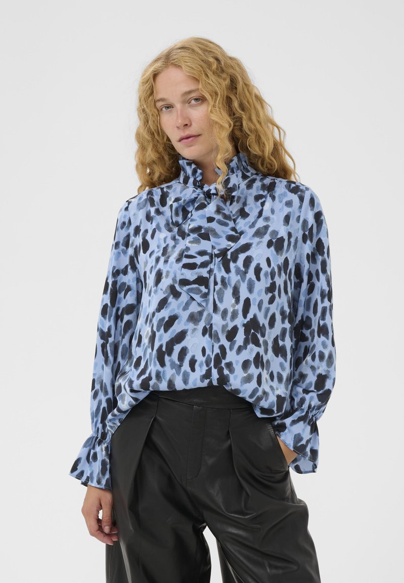 Une femme aux cheveux blonds bouclés porte un blouse bleue à pois noirs avec un nœud au cou, accompagnée d'un pantalon en cuir noir taille haute.