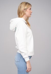 Felpa bianca con cappuccio e cordino, caratterizzata da una vestibilità rilassata e polsini a costine. Abbinata a jeans azzurri, che mostrano un look casual.
