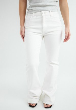 Žena nosi biele rifle s vysokým pásom a rozšírenými nohavicami, svetlosivé zastrčené tričko a čierne sandále s otvorenými prstami na jednoliatom pozadí.