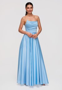 Femme souriante, portant une robe de satin bleu clair, longue jusqu'au sol, sans bretelles, debout avec les mains jointes devant un fond blanc uni.
