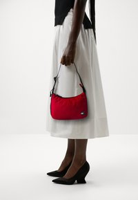 Bolso de mano de nylon rojo con forma curva, cremallera negra y correa ajustable, sostenido por una persona que lleva una falda blanca y tacones negros.