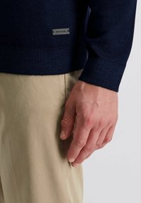 Marineblauwe gebreide trui met geribde manchetten en onderkant, voorzien van een rechthoekig metalen label met de merknaam. Lichtbeige broek is gedeeltelijk zichtbaar.