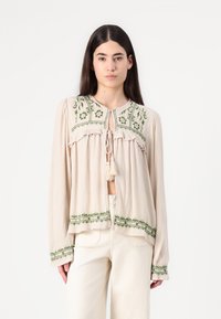 Blouse beige clair avec broderies florales vertes, dotée d'une fermeture à lien, de détails froncés et de manches longues. Confectionnée en tissu texturé.